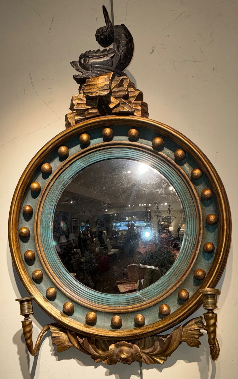 A circular Regency giltwood convex mirror featuring a carved dolphin cresting, ball spherule frame, green inner cove moulding, and lower twin candle sconces.