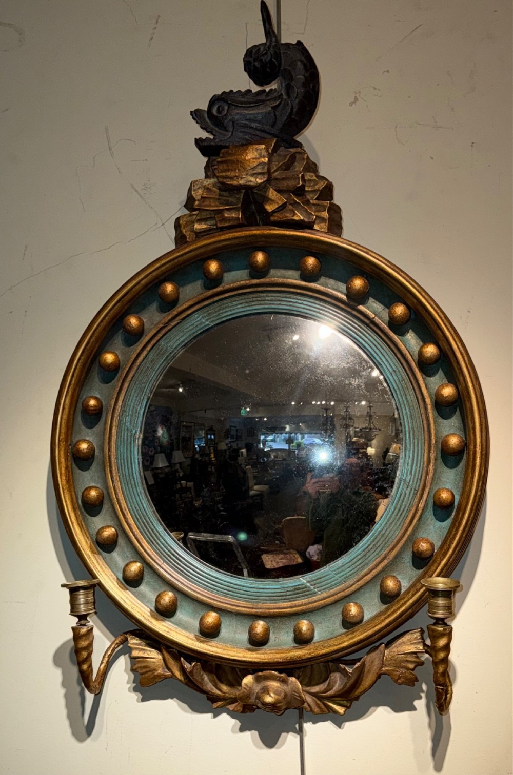 A circular Regency giltwood convex mirror featuring a carved dolphin cresting, ball spherule frame, green inner cove moulding, and lower twin candle sconces. View III