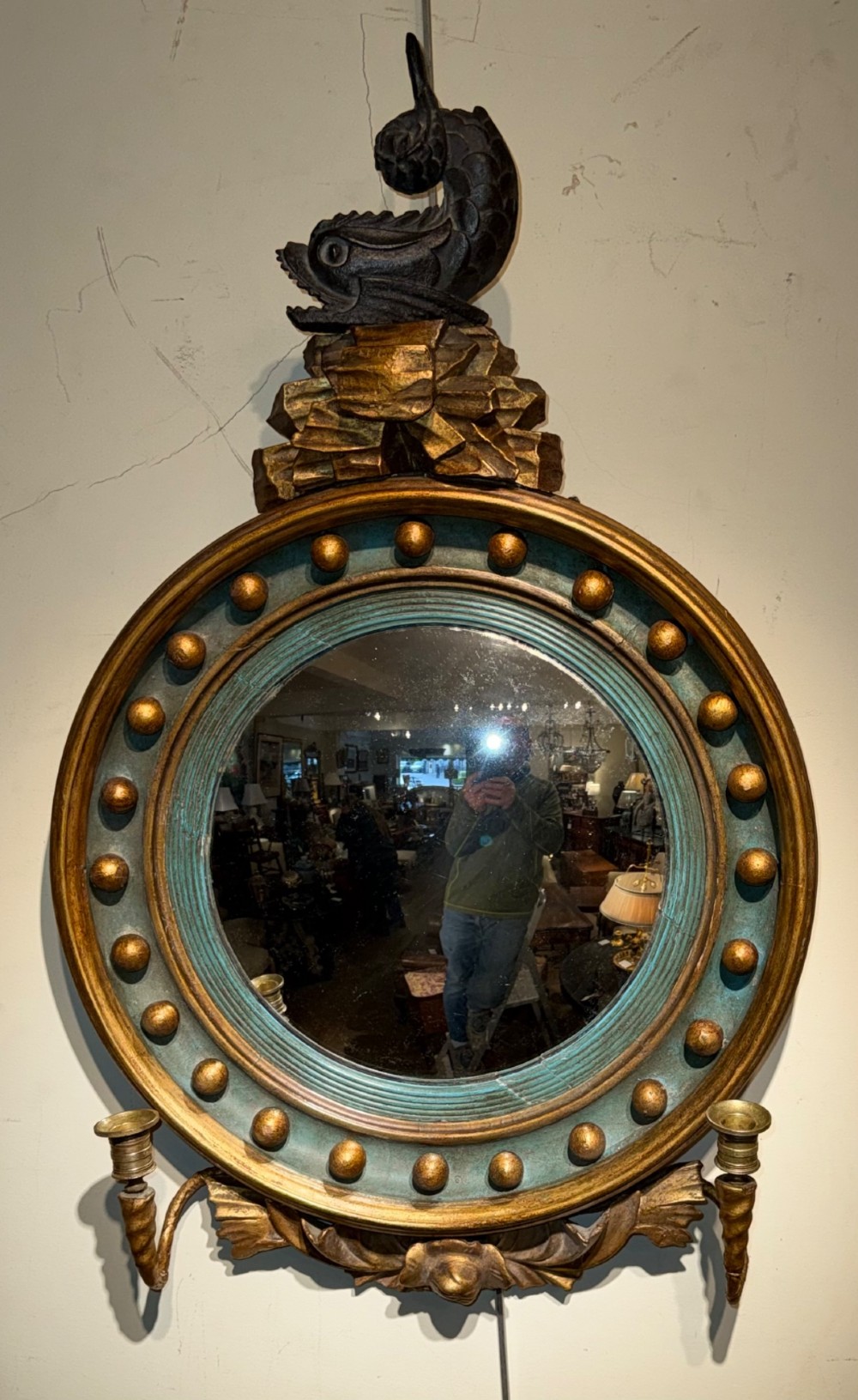 A circular Regency giltwood convex mirror featuring a carved dolphin cresting, ball spherule frame, green inner cove moulding, and lower twin candle sconces. View VI