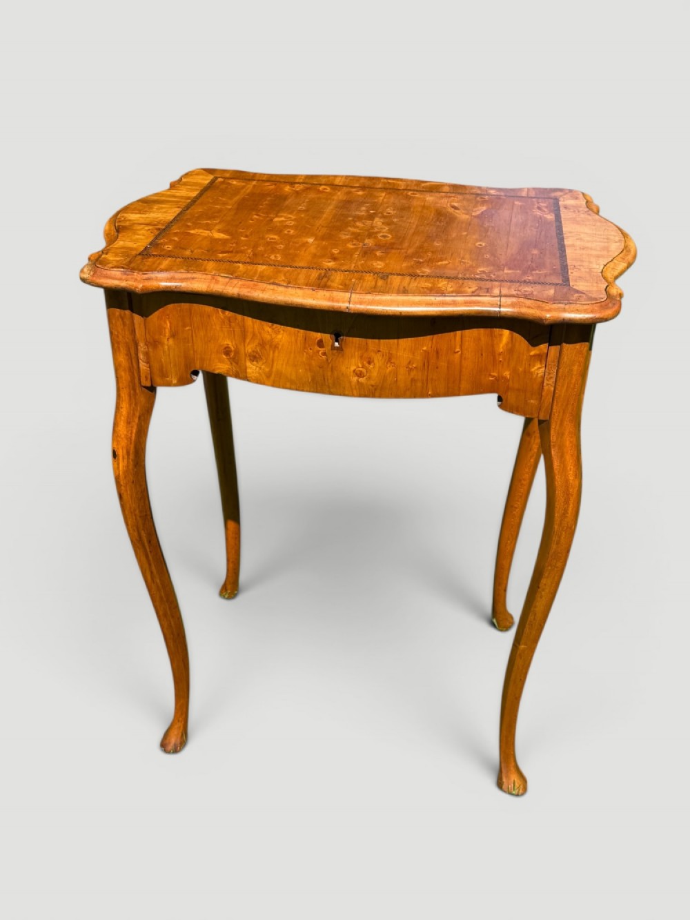 A golden-brown burr wood side table featuring a serpentine-edged top with inlay, a single drawer with a keyhole, and four slender cabriole legs.