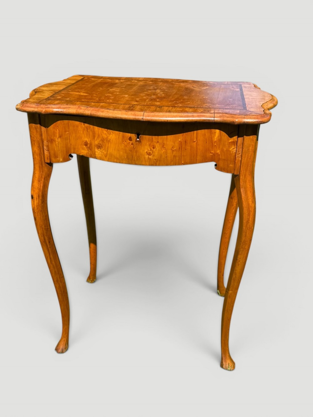A golden-brown burr wood side table featuring a serpentine-edged top with inlay, a single drawer with a keyhole, and four slender cabriole legs. View IV