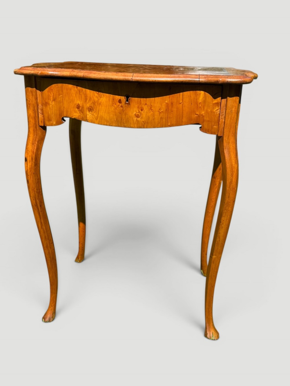 A golden-brown burr wood side table featuring a serpentine-edged top with inlay, a single drawer with a keyhole, and four slender cabriole legs. View VI