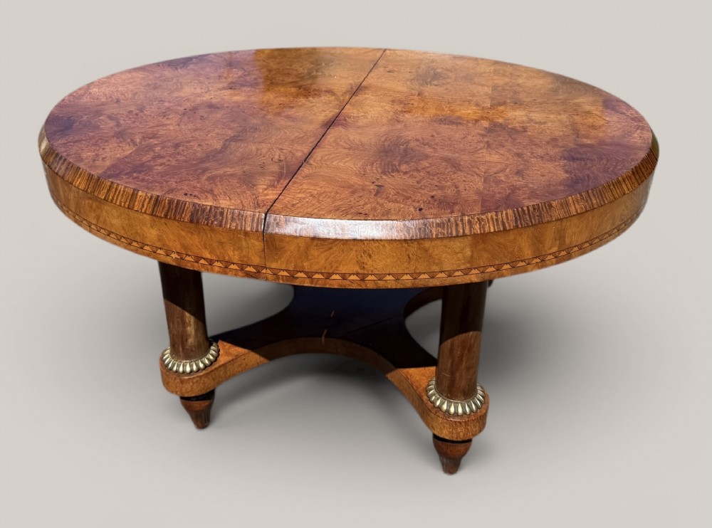 A circular Art Deco style extending dining table made of figured burr walnut veneer with a geometric inlaid apron and a quadruped base.
