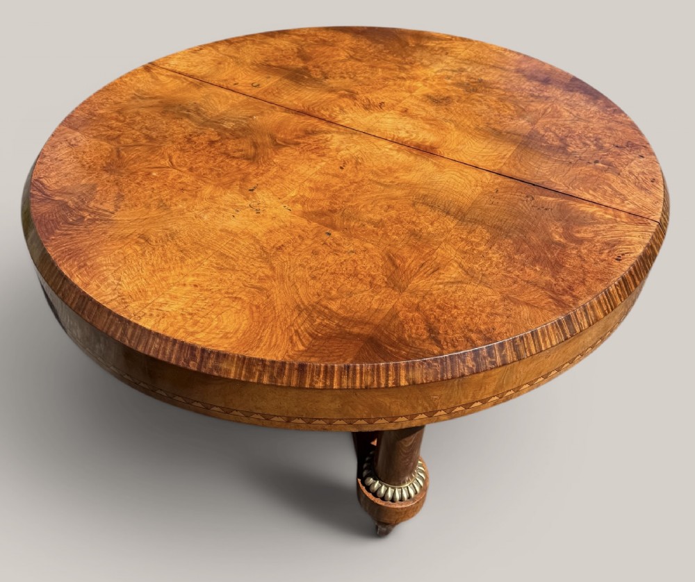 A circular Art Deco style extending dining table made of figured burr walnut veneer with a geometric inlaid apron and a quadruped base. View II