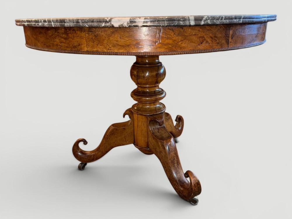 A circular antique gueridon table featuring a grey veined marble top, a figured walnut apron, a turned central pedestal, and scrolled tripod legs on castors.