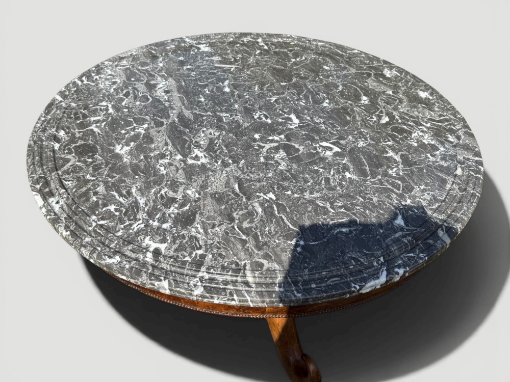 A circular antique gueridon table featuring a grey veined marble top, a figured walnut apron, a turned central pedestal, and scrolled tripod legs on castors. View V