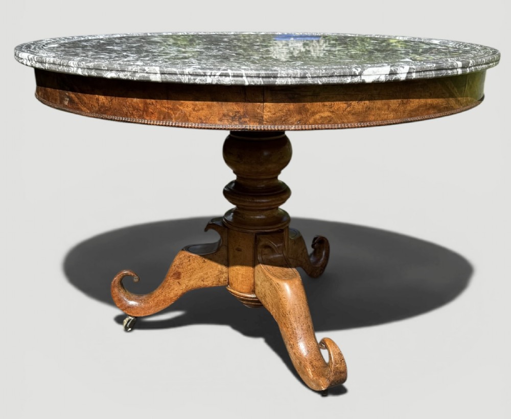 A circular antique gueridon table featuring a grey veined marble top, a figured walnut apron, a turned central pedestal, and scrolled tripod legs on castors. View VI
