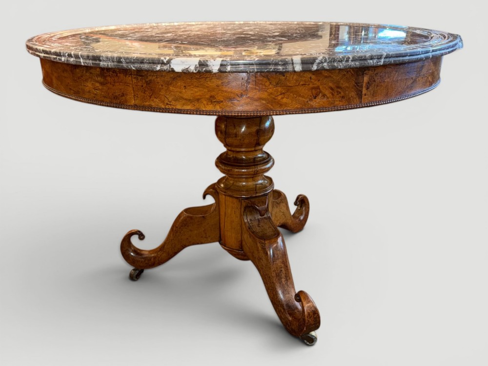 A circular antique gueridon table featuring a grey veined marble top, a figured walnut apron, a turned central pedestal, and scrolled tripod legs on castors. View VII