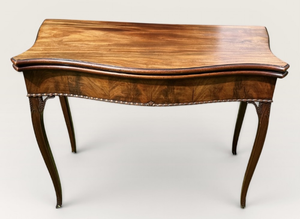 An antique warm brown mahogany card table with a serpentine front, carved cabriole legs, and a hinged fold-over top.