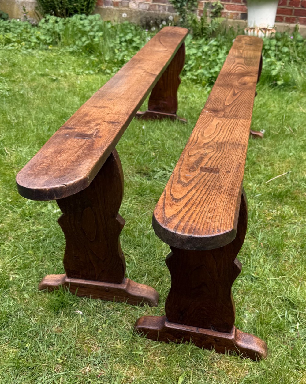 A pair of long French rustic oak benches with shaped trestle legs and smooth rounded seats showing distinct timber grain.