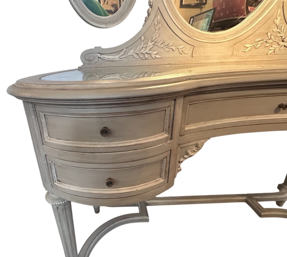 An off-white painted Edwardian kidney-shaped dressing table featuring five drawers, fluted legs, a glass inset top, and an attached triple oval mirror decorated with carved ribbon cresting. View II