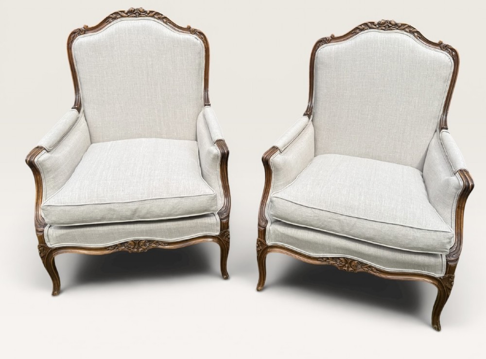 A pair of French Louis XV style Bergere armchairs featuring carved walnut wood frames and neutral light cream linen upholstery.