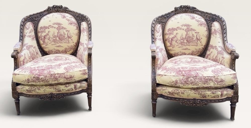A pair of carved wooden French bergere armchairs upholstered in cream and burgundy toile de Jouy patterned fabric.
