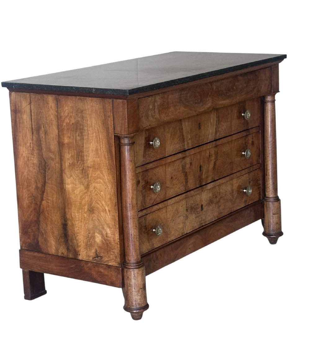 A 19th-century French Charles X walnut commode featuring three lower drawers with round metal pulls, flanked by architectural side columns and topped with a dark marble slab.