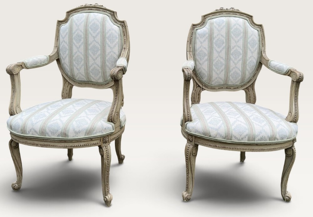 A pair of French armchairs featuring distressed grey painted wooden frames with carved details and striped light blue upholstered seats and backrests.