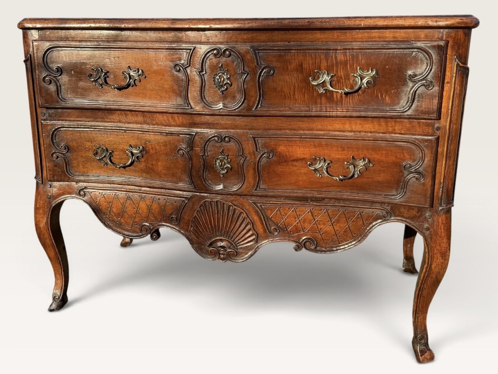An 18th-century French provincial walnut commode featuring a serpentine front, two carved drawers with brass handles, a central carved shell motif on the apron, and cabriole legs.