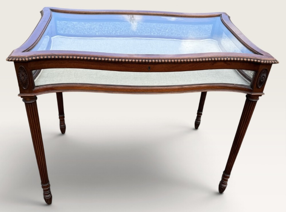 A serpentine-shaped mahogany bijouterie table featuring glazed sides, a glass top, a light fabric-lined interior, and tapered fluted legs.