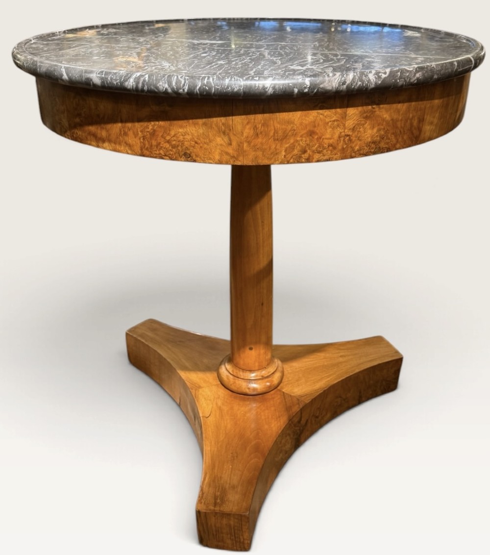 A circular antique centre table featuring a grey veined marble top, burr walnut veneered apron, a central turned column, and a triform platform base.