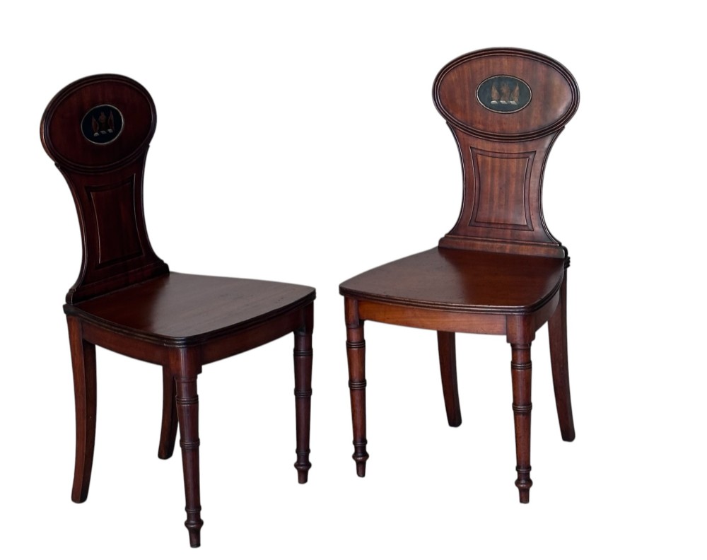A pair of antique dark mahogany hall chairs featuring oval backrests decorated with painted armorial crest medallions, solid timber seats, and turned front legs.