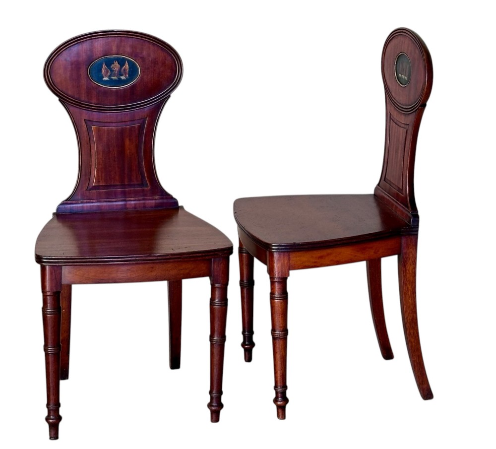 A pair of antique dark mahogany hall chairs featuring oval backrests decorated with painted armorial crest medallions, solid timber seats, and turned front legs. View II