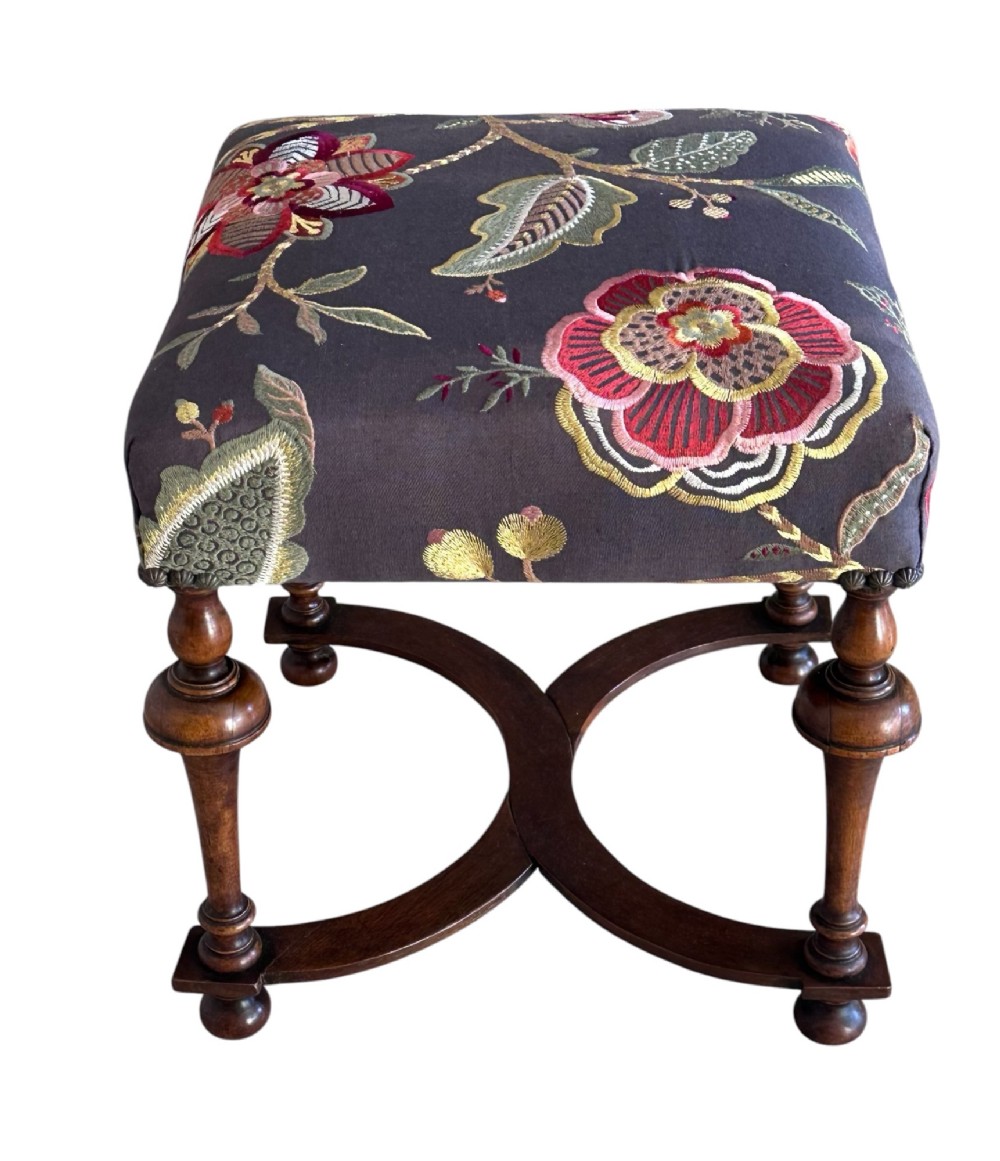 A 19th-century William and Mary style walnut stool featuring turned legs, a curved X-frame stretcher, and dark upholstered top embroidered with colourful floral motifs.