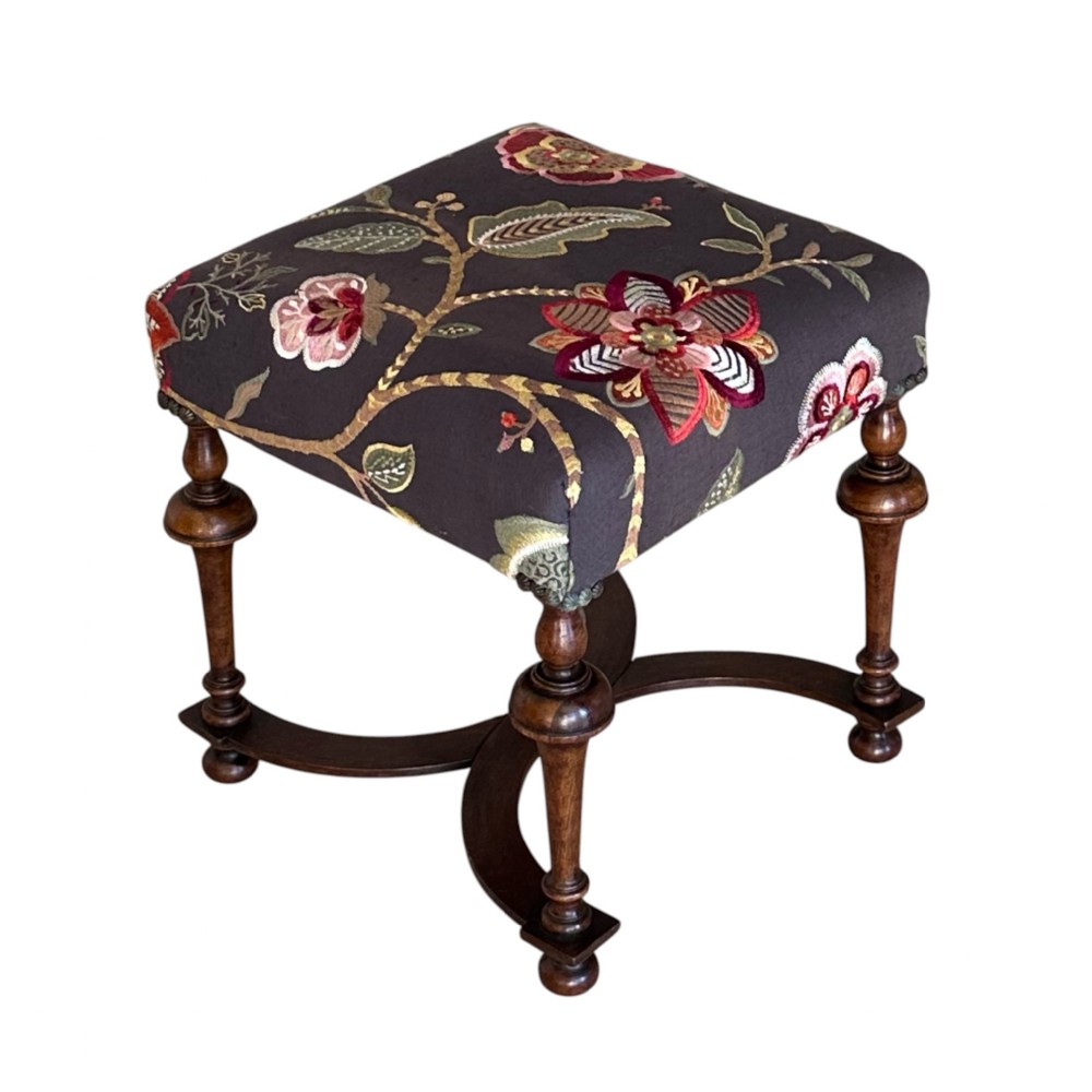 A square 19th-century William and Mary style stool featuring dark turned walnut legs, curved stretchers, and dark textile upholstery embroidered with floral motifs.