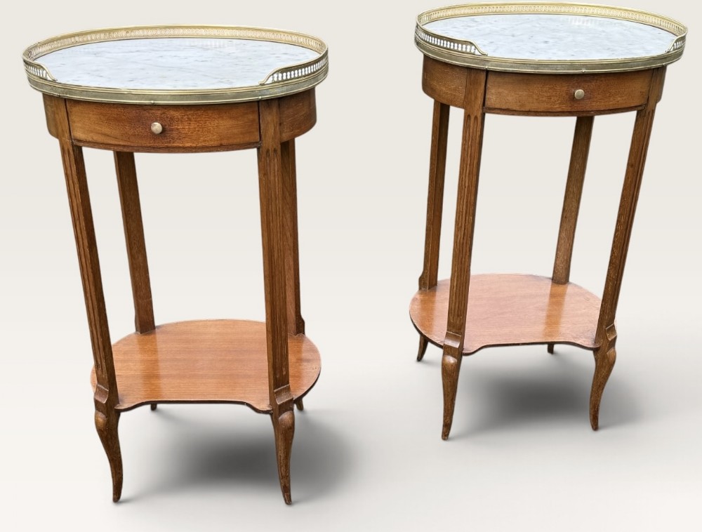 A pair of oval French-style wooden side tables featuring grey-veined marble tops enclosed by pierced brass galleries, single frieze drawers, and lower undertier shelves.
