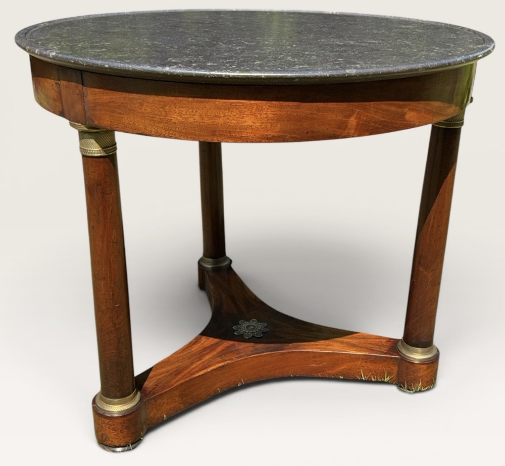 A French Empire style circular centre table with a grey marble top, flame mahogany apron, cylindrical legs with brass mounts, and a tripartite base.