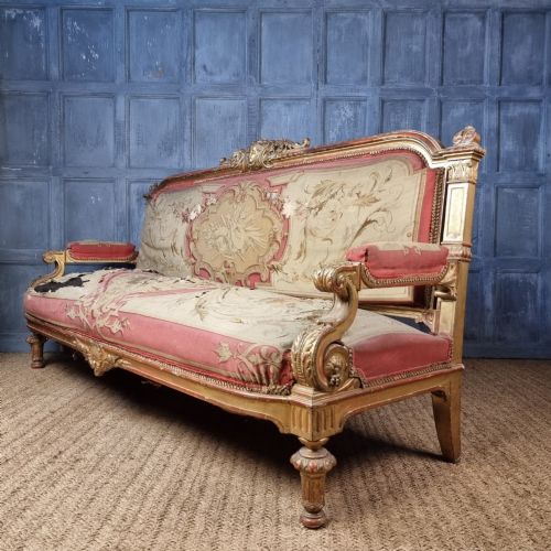 A 19th-century giltwood settee featuring detailed frame carvings, padded armrests, and distressed floral tapestry upholstery. View X