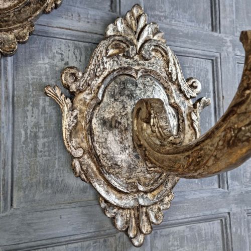 A pair of 17th-century carved wood wall sconces with silvered gilding, featuring ornate cartouche backplates, scrolling arms, and iron prickets. View V