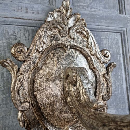 A pair of 17th-century carved wood wall sconces with silvered gilding, featuring ornate cartouche backplates, scrolling arms, and iron prickets. View X