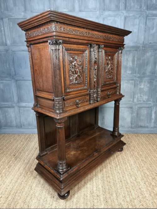 A dark carved oak cupboard featuring two relief-carved doors with figure motifs, upper drawers with ring handles, fluted columns, and an open lower tier. View X