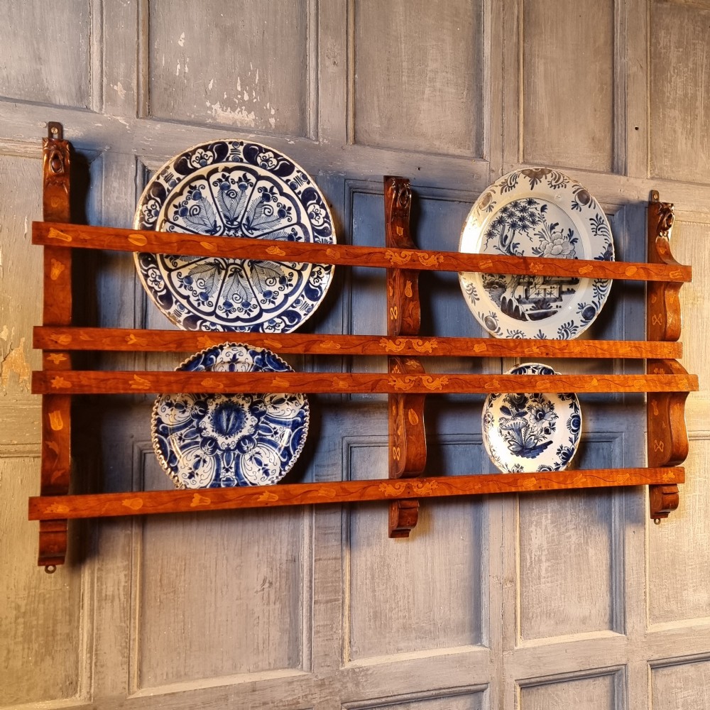 A wall-mounted 19th-century wooden delft rack featuring horizontal plate rails decorated with floral marquetry inlays.