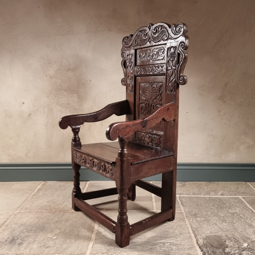 A dark carved oak seventeenth-century wainscot armchair featuring a panelled backrest, scrolling ears, turned front legs, and square stretchers. View XIII