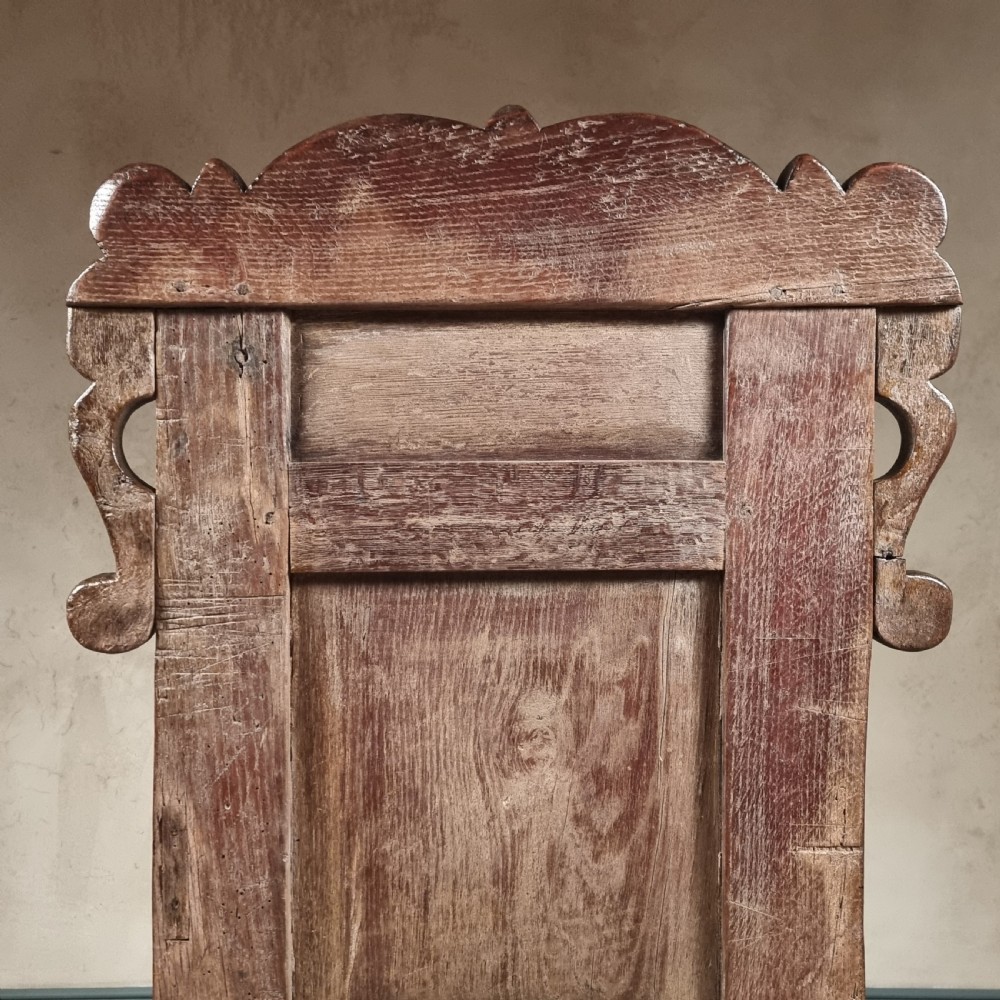 A dark carved oak seventeenth-century wainscot armchair featuring a panelled backrest, scrolling ears, turned front legs, and square stretchers. View XV