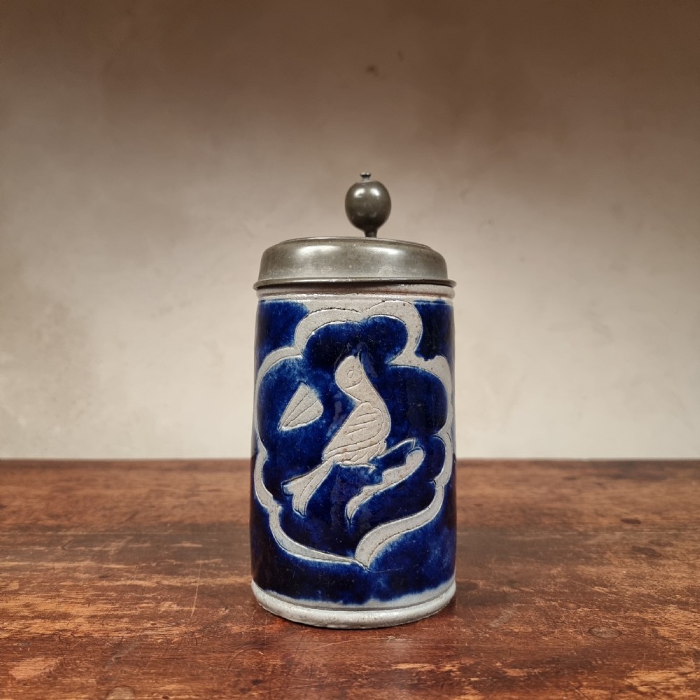 An 18th-century cylindrical Westerwald grey salt-glazed stoneware tankard with cobalt blue glaze, featuring an incised bird motif inside a lobed panel and a hinged pewter lid.