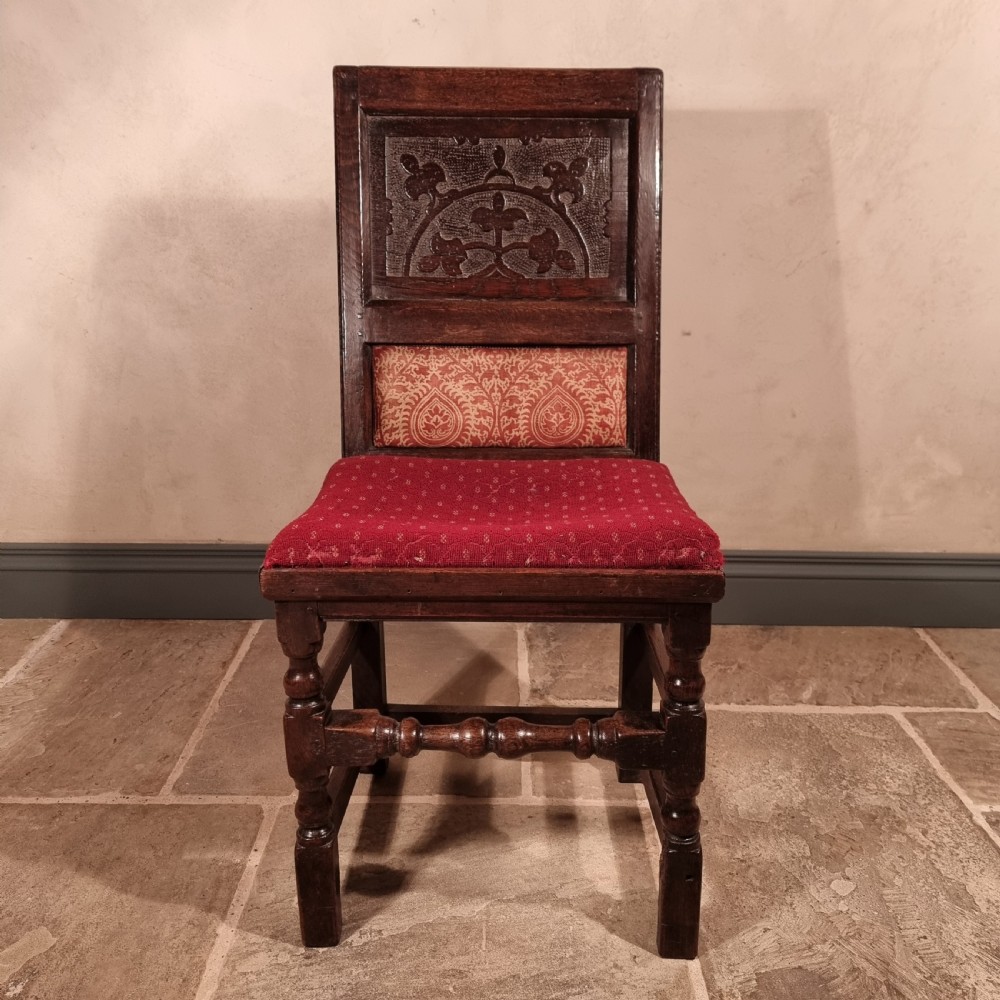 A dark carved oak side chair with an arched floral back panel, red upholstered lumbar cushion, red seat, and turned front legs. View V
