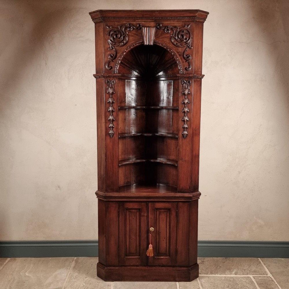 A tall dark mahogany corner cupboard featuring an open carved arched alcove with shaped shelves above a two-door lower cupboard.