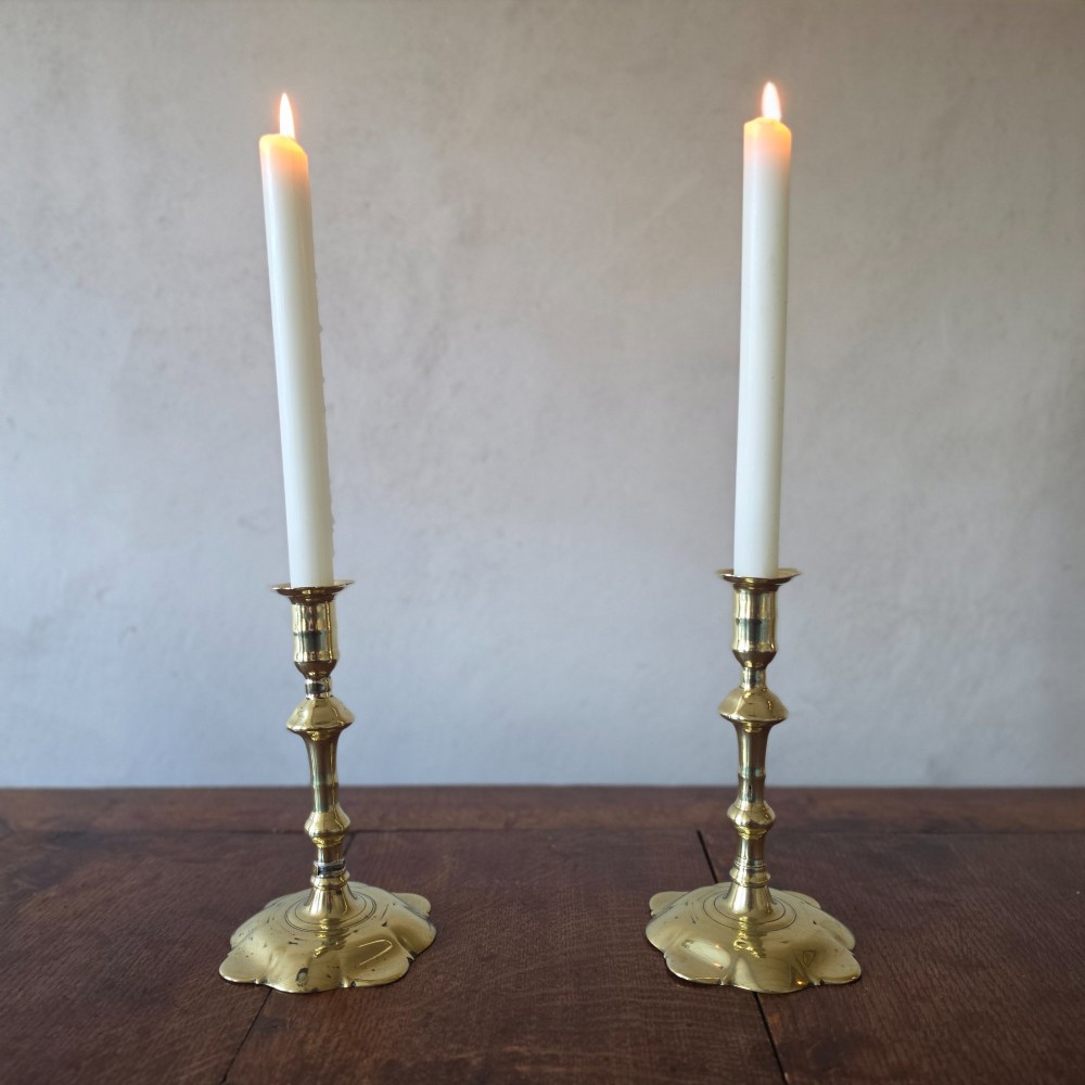 A pair of 18th-century brass candlesticks featuring turned baluster stems and scalloped petal bases, holding lit white taper candles.