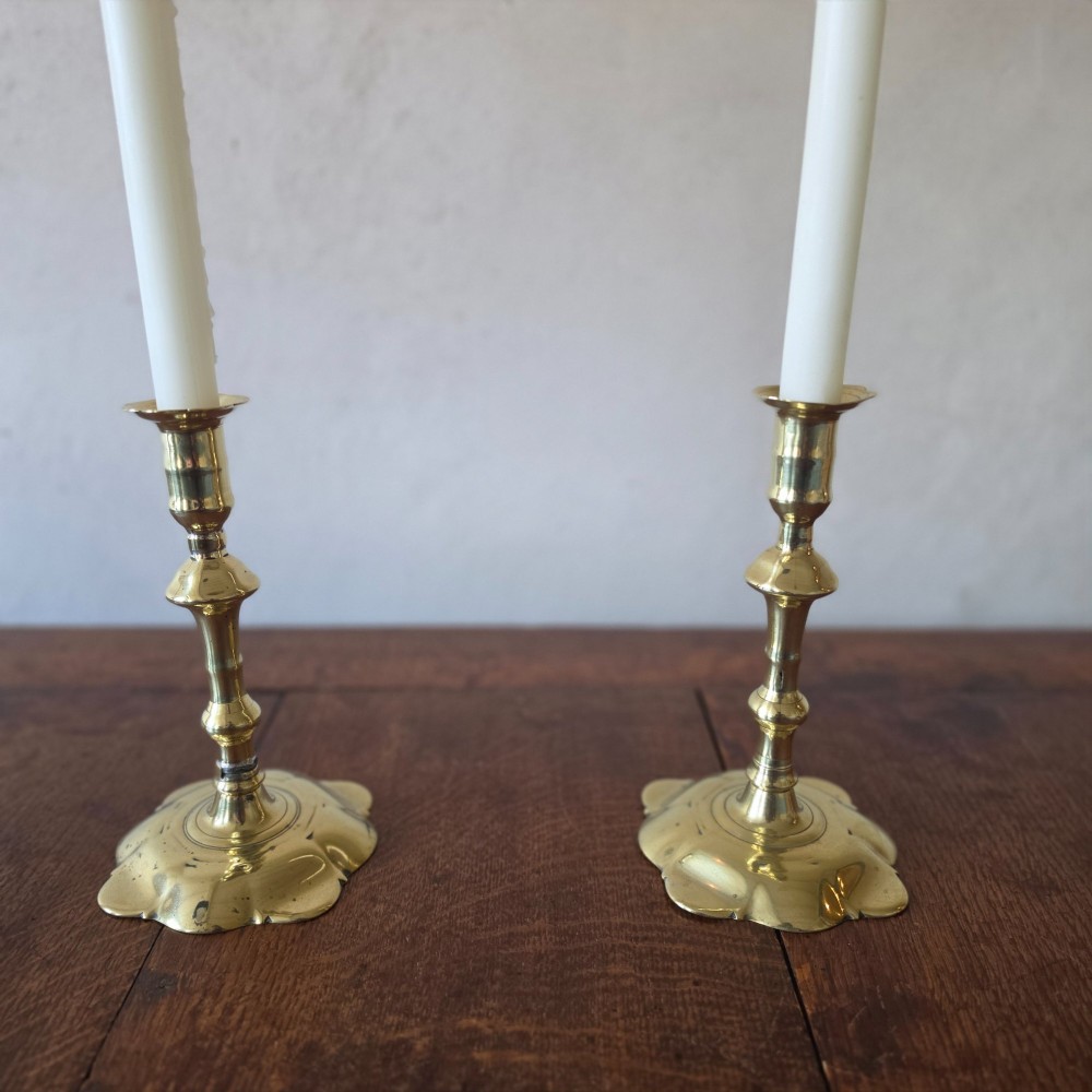 A pair of 18th-century brass candlesticks featuring turned baluster stems and scalloped petal bases, holding lit white taper candles. View II