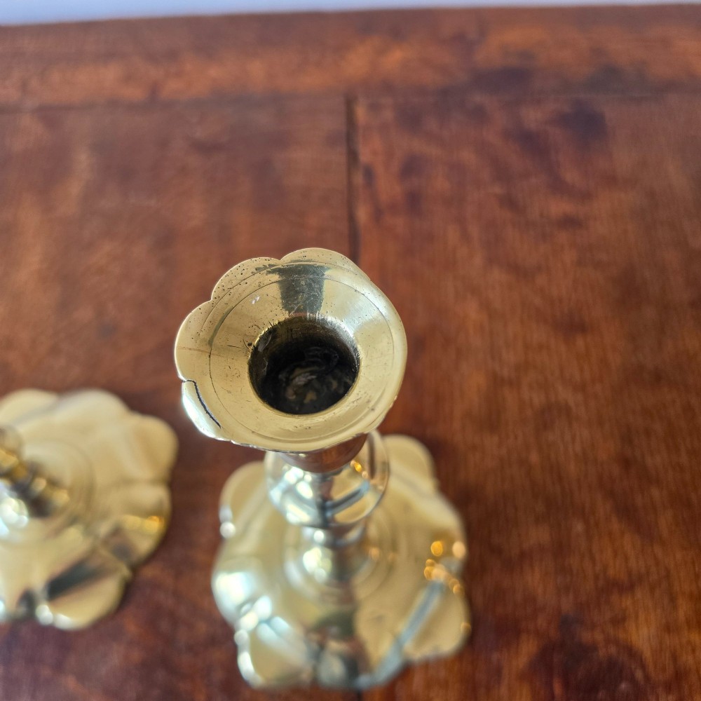 A pair of 18th-century brass candlesticks featuring turned baluster stems and scalloped petal bases, holding lit white taper candles. View V