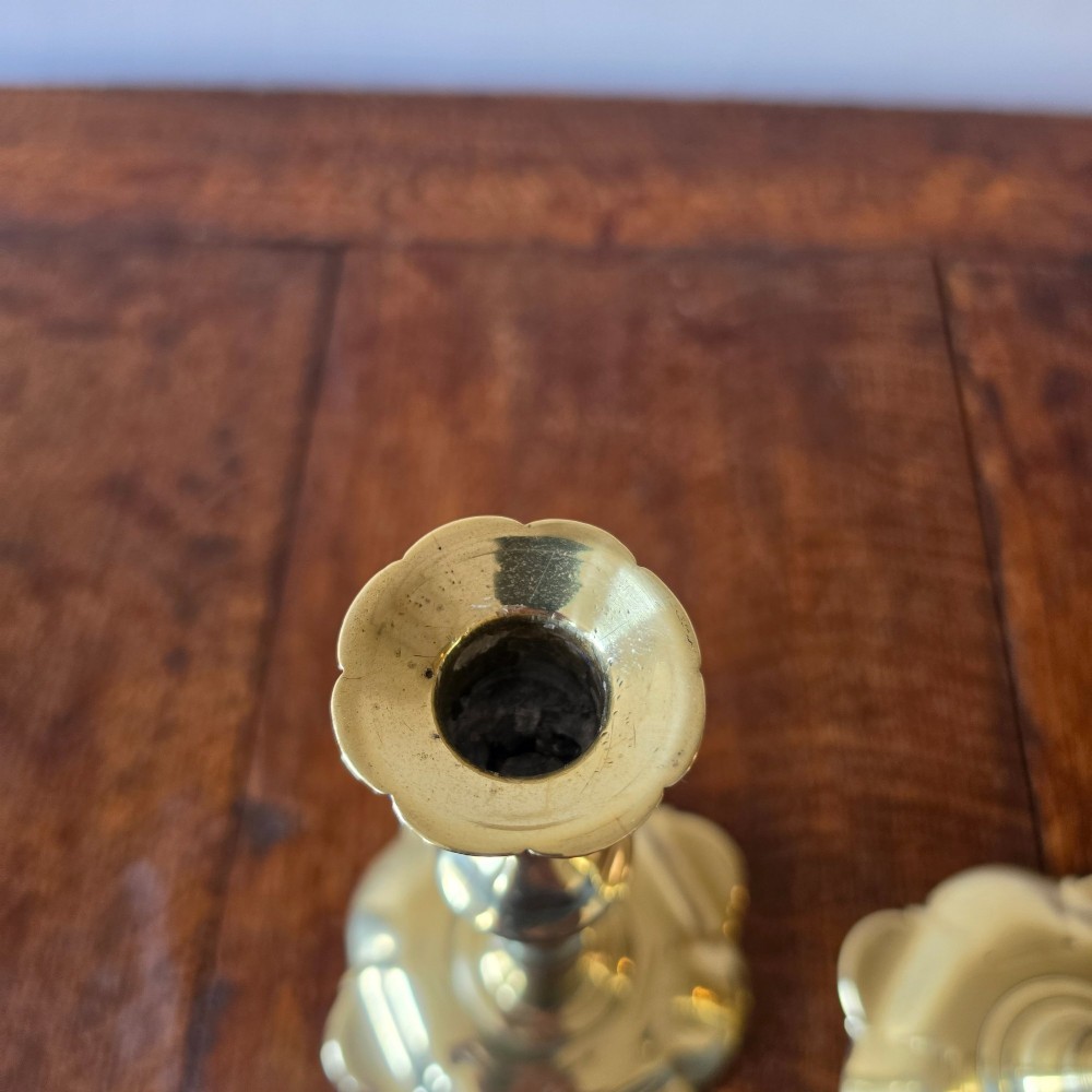 A pair of 18th-century brass candlesticks featuring turned baluster stems and scalloped petal bases, holding lit white taper candles. View VI