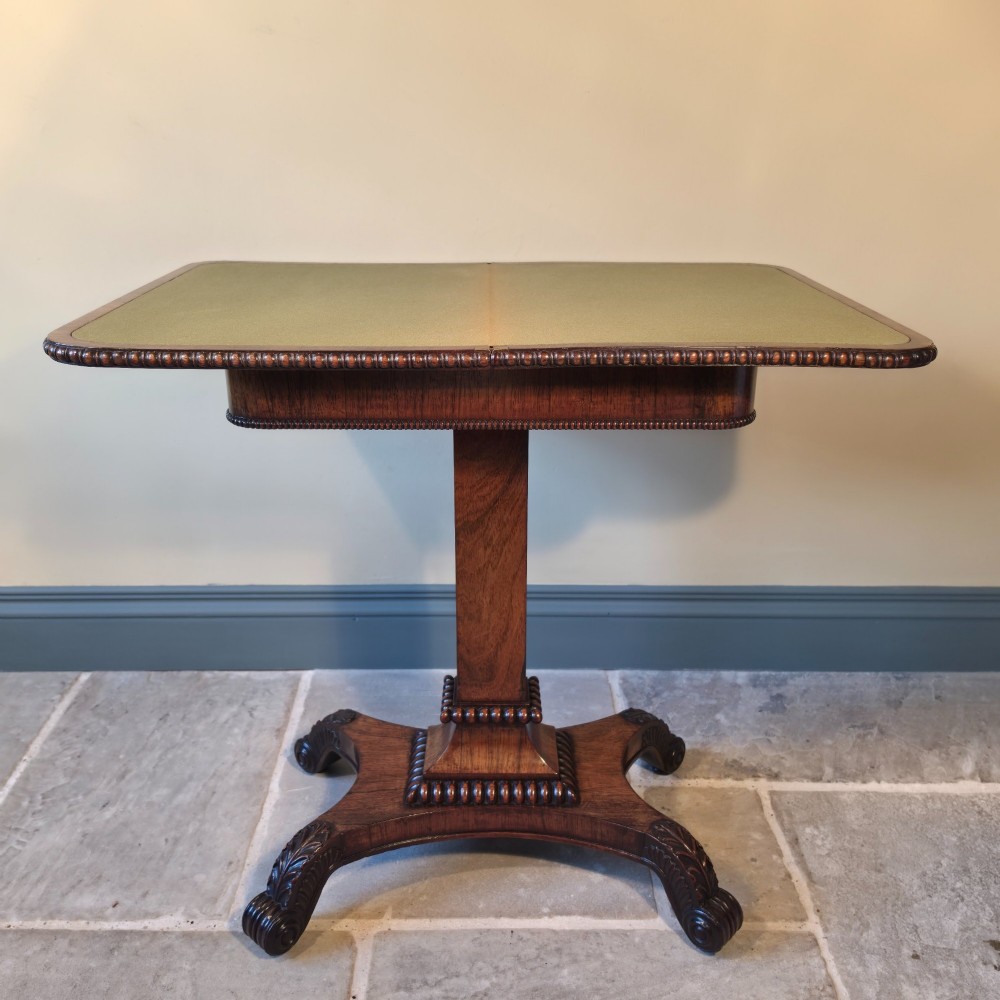 A William IV rosewood pedestal card table with a green baize playing surface, bead moulding, and carved scroll feet.