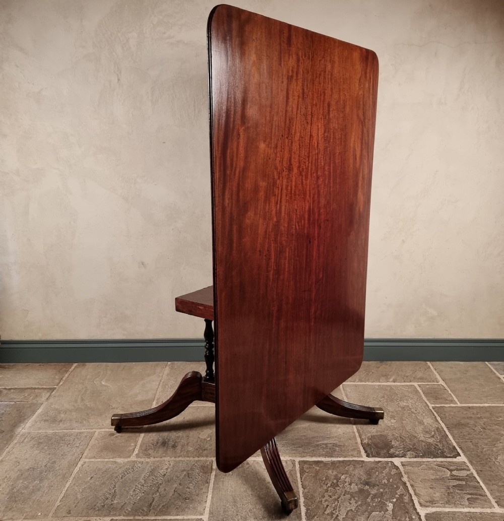A Regency mahogany tilt-top dining table shown secured vertically on a central pedestal with reeded swept legs and brass castors.