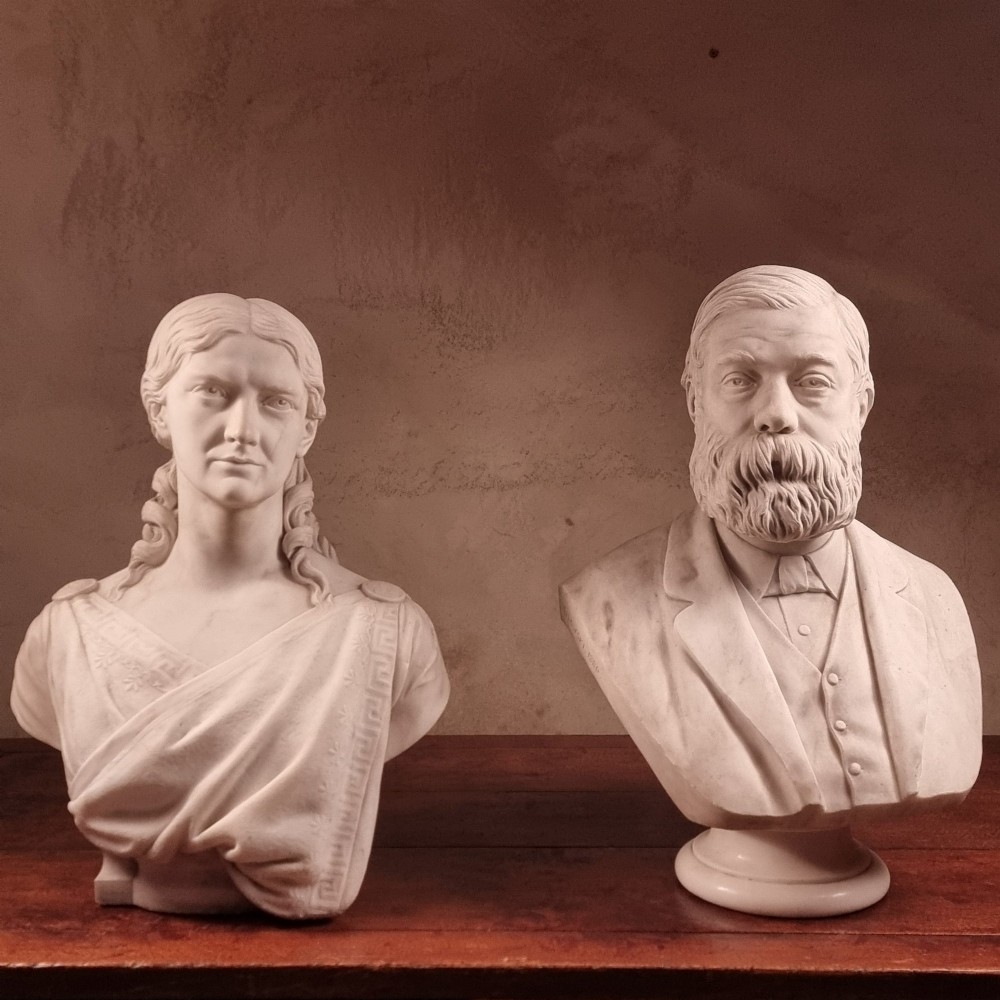 A pair of carved white marble portrait busts depicting a classical female figure on the left and a bearded man in Victorian attire on the right.