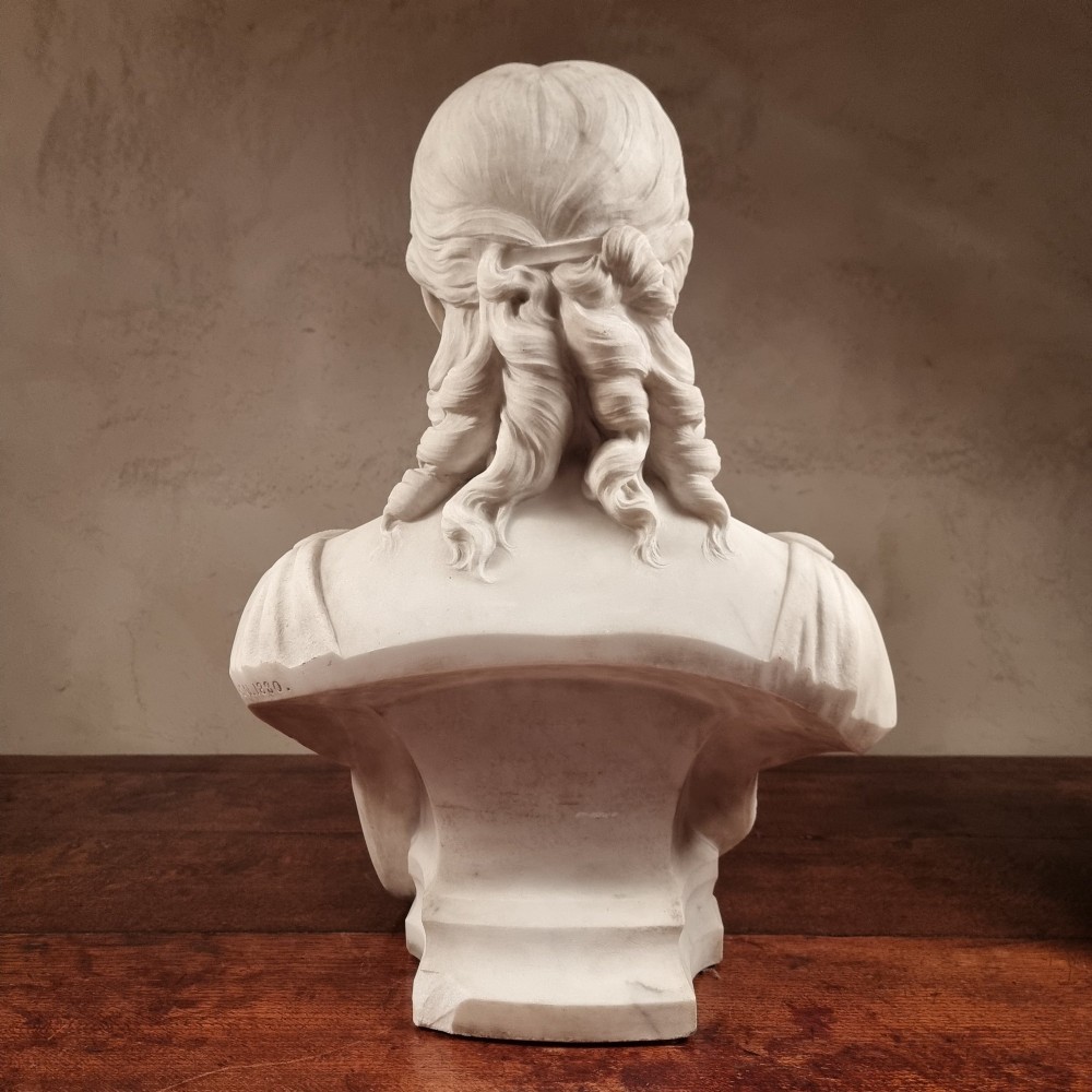 A pair of carved white marble portrait busts depicting a classical female figure on the left and a bearded man in Victorian attire on the right. View X