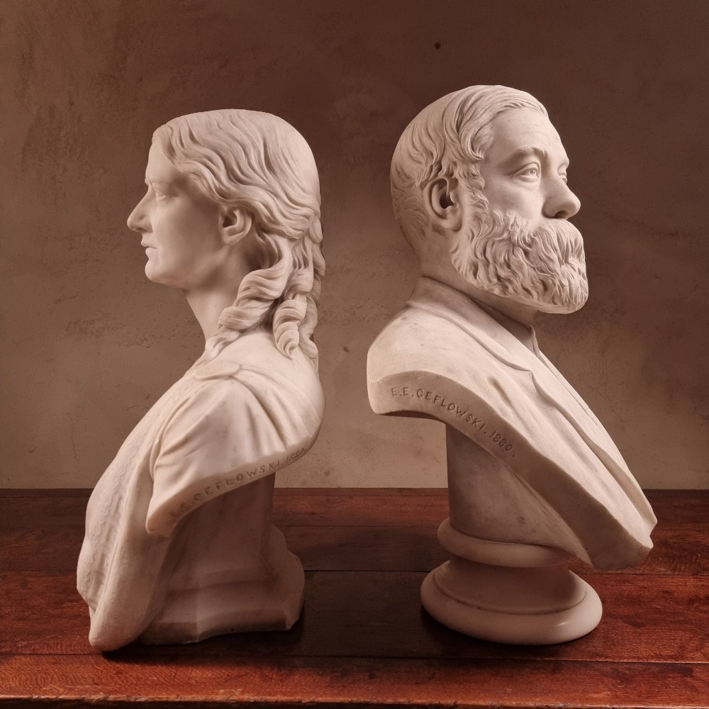 A pair of carved white marble portrait busts depicting a classical female figure on the left and a bearded man in Victorian attire on the right. View XV