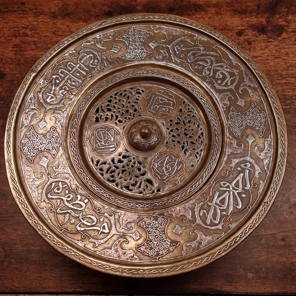 A circular 19th-century Islamic brass wash bowl featuring intricate Arabic calligraphic script, geometric arabesque patterns, pierced metalwork, and silver and copper inlay.