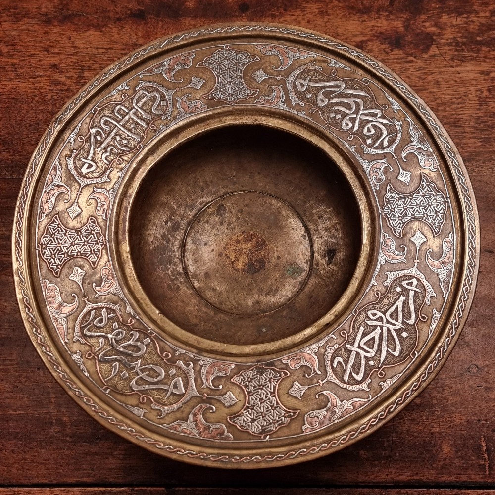 A circular 19th-century Islamic brass wash bowl featuring intricate Arabic calligraphic script, geometric arabesque patterns, pierced metalwork, and silver and copper inlay. View II