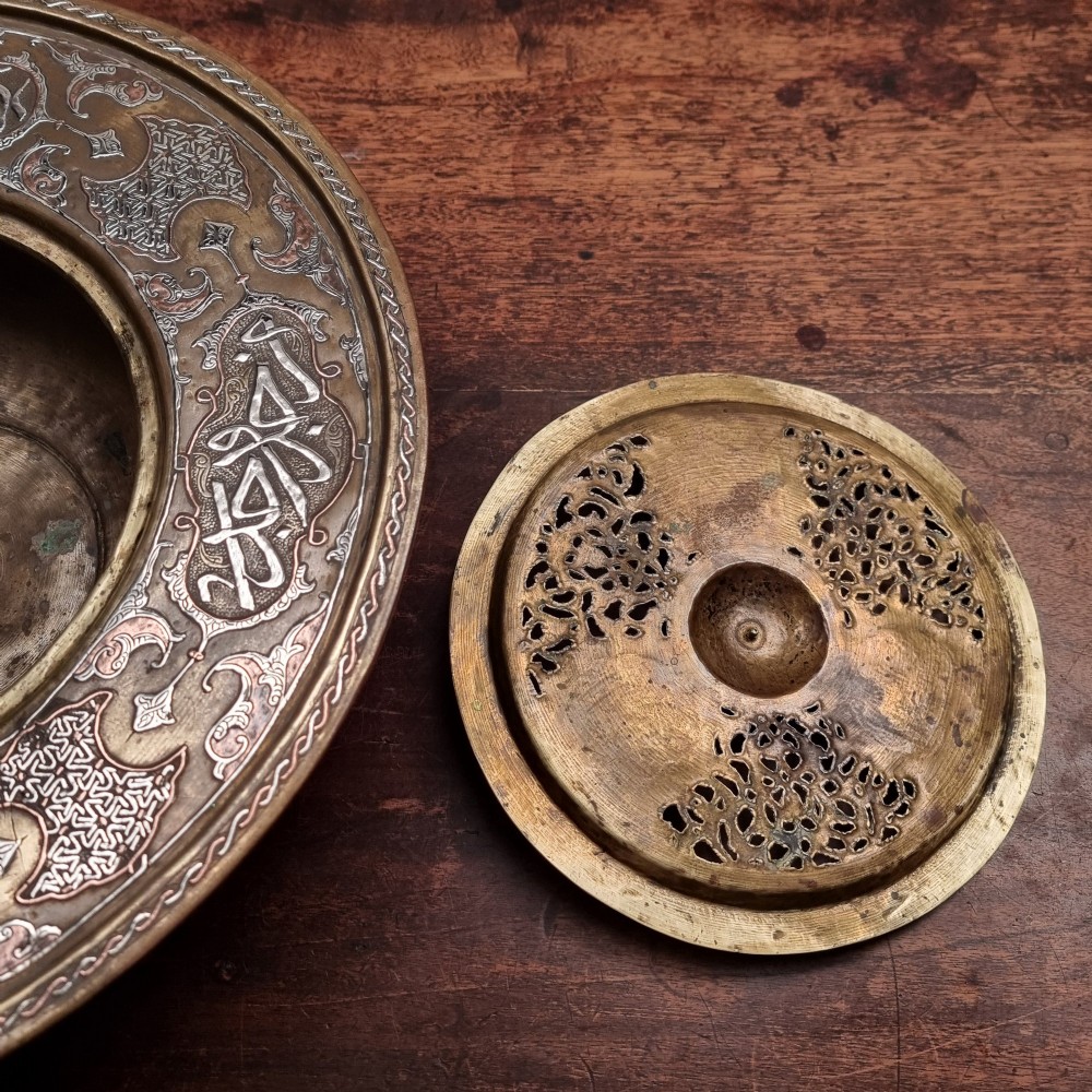 A circular 19th-century Islamic brass wash bowl featuring intricate Arabic calligraphic script, geometric arabesque patterns, pierced metalwork, and silver and copper inlay. View III